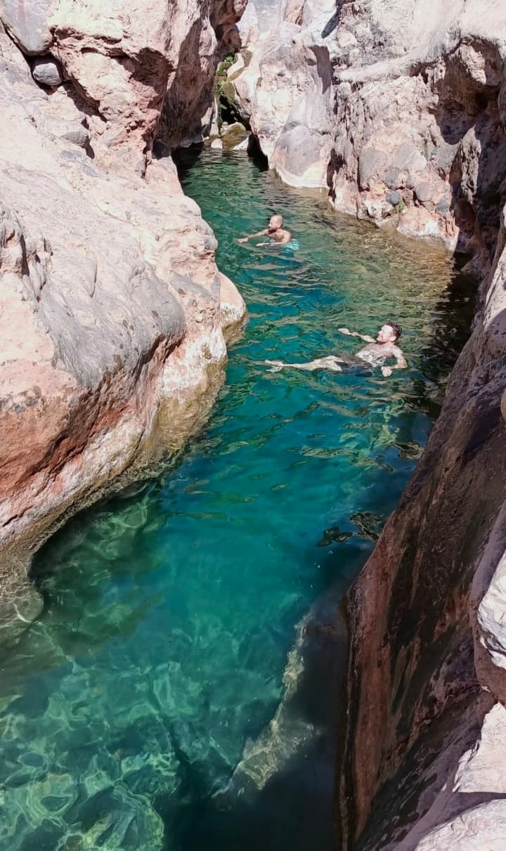 A narrow turquoise pool between dark canyon walls with two people swimming